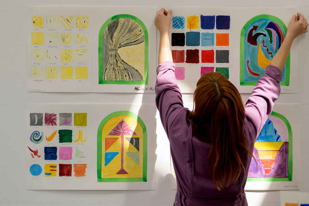 A photograph taken perhaps in a workshop or studio. Four large pieces of paper are hanging on a wall in a square formation, three of which are fully visible, with the one bottom right partially obscured by a white woman with long hair, wearing a mauve top, who is fixing the top right picture to the wall. Each page of paper has 16 small boxes on the left, in rows of 4, which seem to be colour or style tests, and an arched window-shape on the right. Each of the small boxes is labelled with a different word, including ‘grief’, ‘beauty’, ‘resilience’, and ‘patience’, and each is illustrated differently, and painted with different colours. Many of the words are the same between the 16 boxes of the 3 visible pictures on the wall, but each is illustrated differently, and each of the window shapes next to them contains a very different drawing made up of colours and patterns that clearly draw on each picture’s 16 boxes.