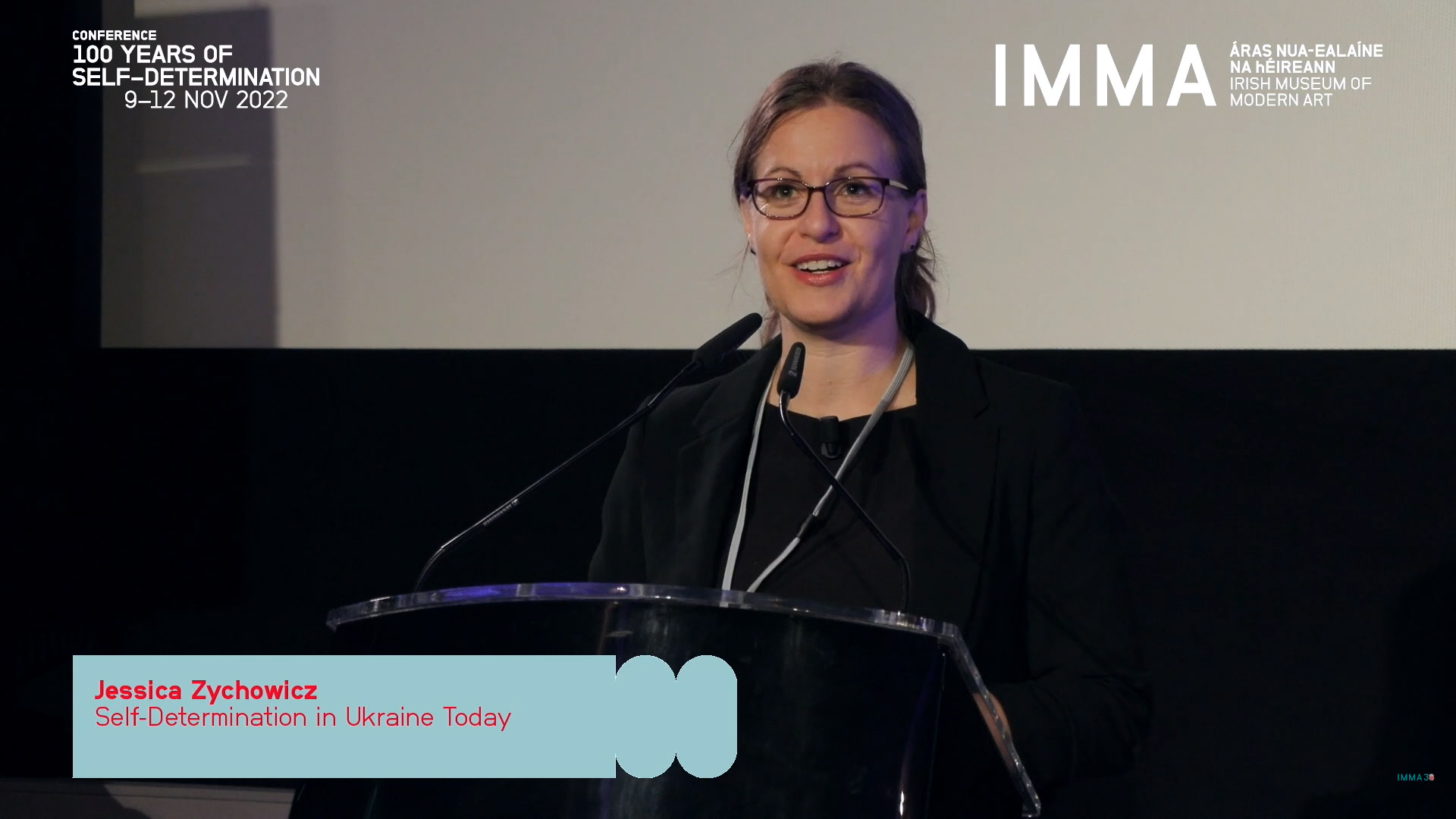 A screenshot from what appears to be a broadcast of a conference. A white woman with brown hair, visible only from the waist up, wearing glasses and dressed in black, is standing at a podium talking into two microphones. She is smiling. Behind her the bottom of a large projector screen is visible. Overlaid in the top left are the words ‘CONFERENCE, 100 YEARS OF SELF-DETERMINATION, 9-12 NOV 2022’, and in the top right it reads ‘IMMA ÁRAS NUA-EALAÍNE NA hÉIREANN; IRISH MUSEUM OF MODERN ART’. In a blue box overlying the photograph on the bottom left is written: ‘Jessica Zychowicz, Self-Determination in Ukraine Today’.