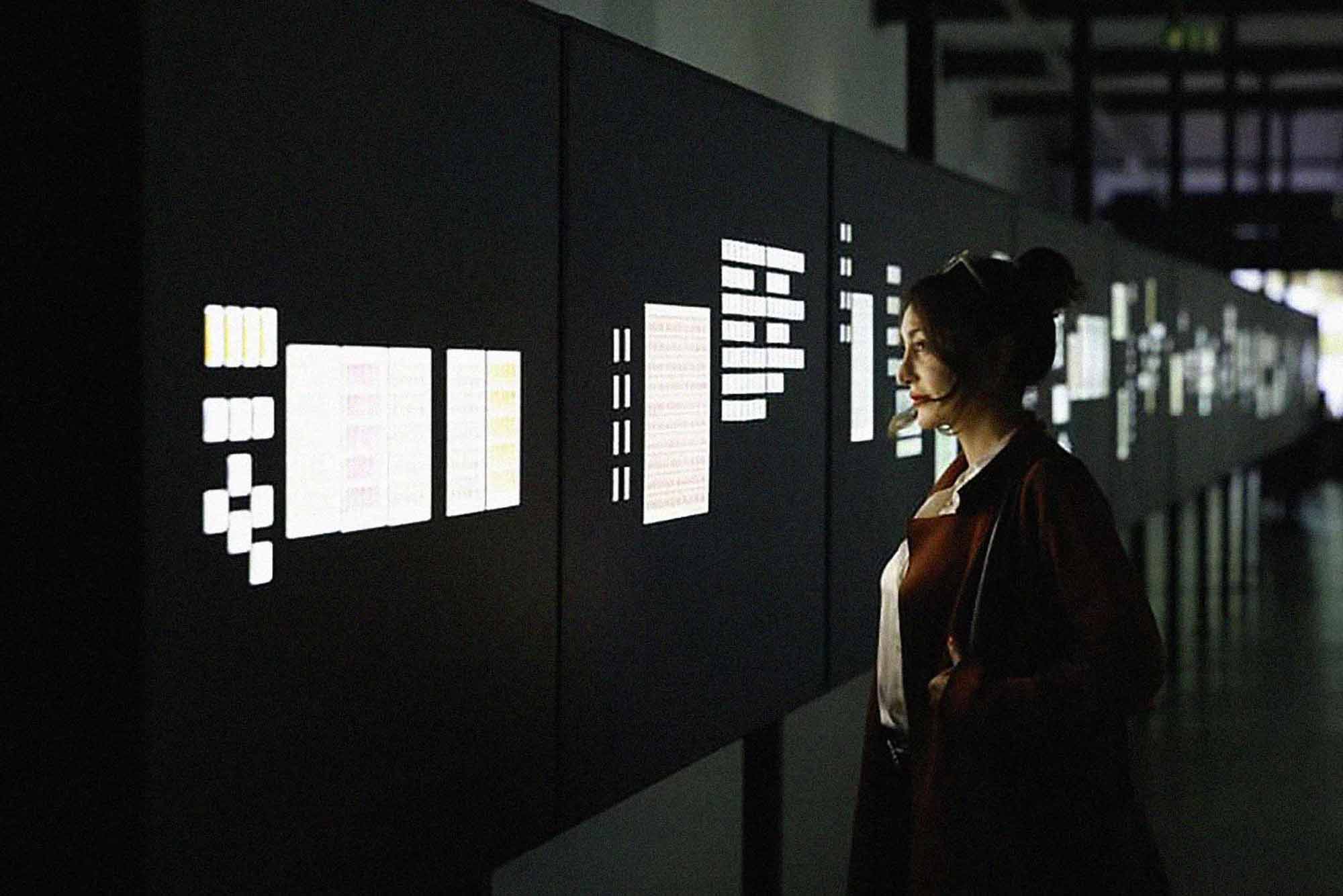 Interior installation photograph inside a long dark corridor, with display panels showing illuminated stamps. A woman stands is looking at the display.