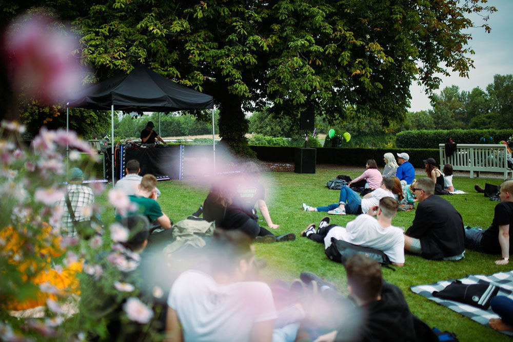 A photograph of a group of people sitting and lying on grass can be seen through out-of-focus flowering shrubs in the foreground. They are looking to the viewer’s left, at a DJ behind a mixing desk under a black awning supported by metal poles, which has been set up under a tree. He is holding a microphone and wearing a cap, and his head is bowed so that his face can’t be made out. To the right of the tree there is a large speaker on the ground. A couple of balloons, one green and one yellow, can be seen in the background behind a hedge. To the right is a pale green wooden bench facing away from the camera, and through the bars of the back someone with rainbow coloured hair can be seen sitting on it.