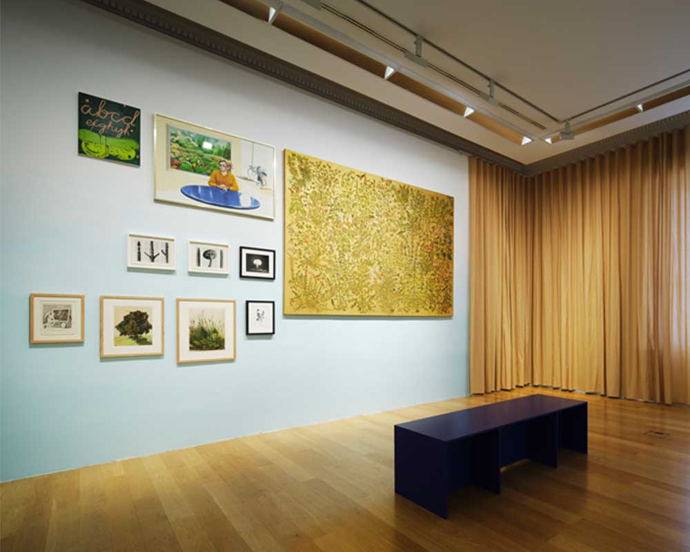 This is a photograph of a gallery room at IMMA, with a light-coloured, polished wooden floor, hung with a selection of pieces from The Narrow Gate exhibition. Rails with lights run along the edges of the ceiling. To the right is a large window. A light, floor-length, golden coloured curtain covers the left-hand side of the window, and continues into the corner of the room, meeting a similar curtain which could be drawn to cover the wall perpendicular to the window, but is drawn back. The wall is a pale blue, and is hung with, from right to left, a large golden coloured landscape canvas or board covered with botanical drawings mostly in a muted green but with touches of blue, white, red and black; a smaller framed painting hung level with the top of the larger canvas, depicts a white man in an apricot coloured shirt sitting at a shiny, round, blue table with an electric fan on a shelf to his left and a painting of a garden with flowers behind him; further to the left, but hung at the same height is another, smaller, landscape canvas or board. This has a black background with two green, curved shapes tied together taking up the bottom half, and ‘abcd’ in large cursive orange writing, with ‘efghijk’ in slightly smaller but similar writing, underneath. Below that is a row of three much smaller black and white images, most likely photographs. The first are in white frames and contain three botanical close up images of stems and a mushroom, juxtaposed starkly with the third photograph on the right which has a black frame and shows a mushroom cloud after a nuclear explosion. Along the bottom below all but the largest canvas are four images of varying sizes, which all appear to be prints. The first shows a sculpture of a seated winged figure to the left in front of a blackboard on an easel with a globe below it; the second shows a row of trees receding from the viewer; the third depicts grasses and bullrushes; and the last what might be botanicals tied in a bunch. There is a rectangular black bench seat a metre or so from the wall.